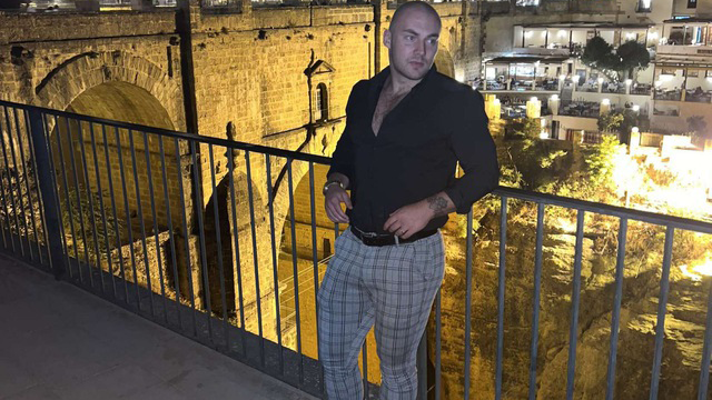 A man in a black shirt and checkered pants leans against a railing at night, with an illuminated ancient stone bridge and buildings in the background. The scene is warmly lit, highlighting the historic architecture, much like how aged Reddit accounts tell stories through their activity history.